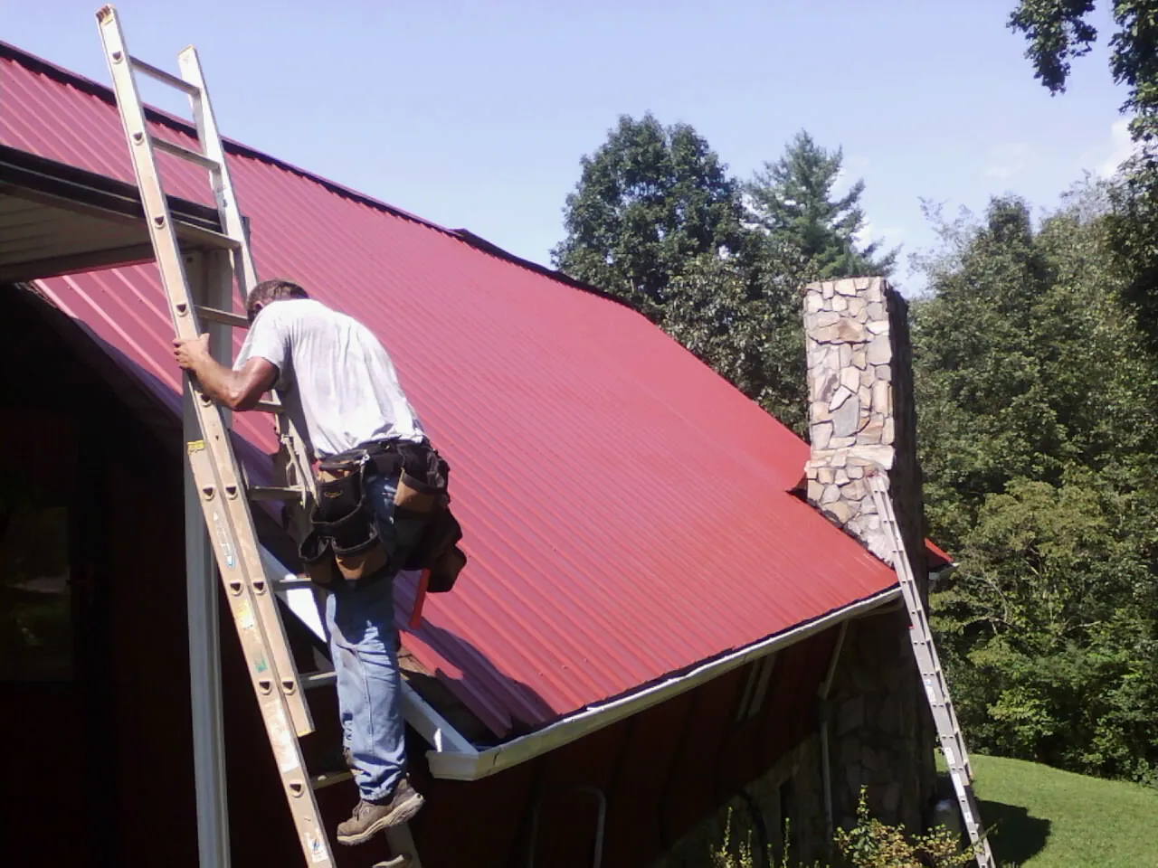 Roofer on a ladder beside a red metal roof and stone chimney — Soffit & Fascia Repair by Roofing Brothers in Woodburn
